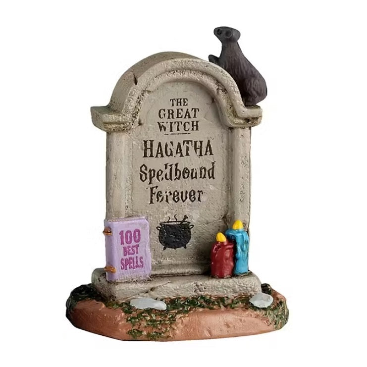 This tombstone reads "THE GREAT WITCH. HAGATHA SPELLBOUND FOREVER." On top of the tombstone, there's a rat. Resting up against the tombstone is a pink book that reads "100 BEST SPELLS" and there are two lit candles: one is red and one is blue.