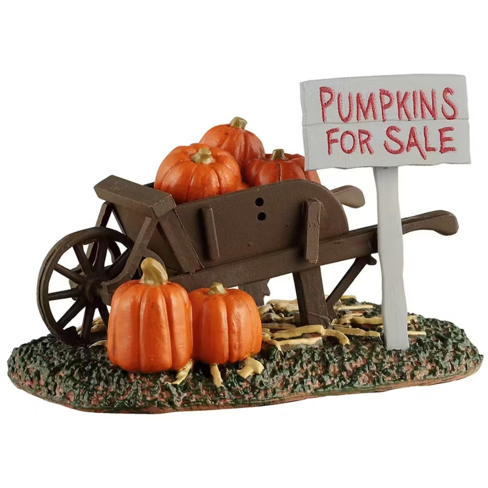 This is a Lemax Spooky Town piece designed to look like a brown wheelbarrow with pumpkins in it. There are also pumpkins underneath it and a sign next to it all that reads "PUMPKINS FOR SALE."