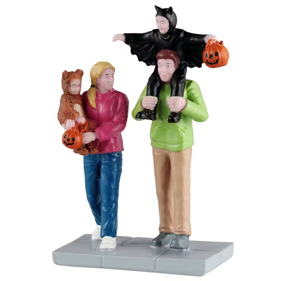 This is a Lemax Spooky Town figurine that is designed to look like two parents taking their kids trick-or-treating. Mom is wearing a pink jacket and blue jeans. Dad is wearing a lime green jacket and khaki pants. Mom is holding a baby that's wearing a bear costume. Dad has a child sitting on his shoulders, and that child is dressed up like a bat. Both kids are holding trick-or-treat bags that look like jack-o-lanterns.