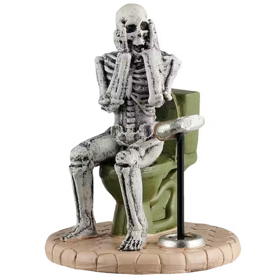 This is a Lemax Spooky Town figurine that's designed to look like a skeleton sitting on a green toilet. With his hands on his face, shocked, he's looking at the toilet paper holder and realizing there's no toilet paper left.