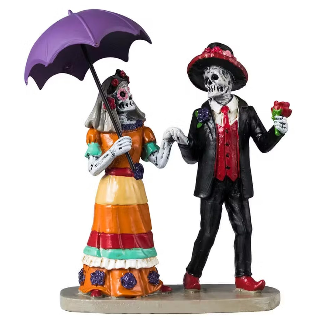 This image shows a Lemax Spooky Town figurine designed to look like two skeletons dressed up for Dia de los Muertos. The skeleton on the left is a woman wearing an orange, teal, red, yellow and purple striped dress and is holding a purple umbrella. The skeleton of the right is a man wearing a black suit with a red vest and red shoes. He's holding a red rose in his hand.