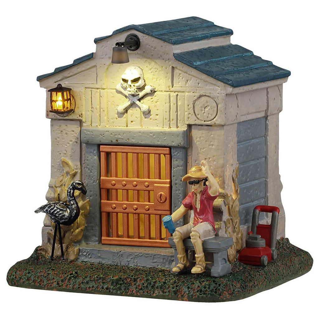 This is a Lemax Spooky Town building designed to look like a mausoleum. There's a man sitting outside on a bench with a lawn mower behind him. There's a black flamingo statue next to the front gate/door. There's a skull and cross bones above the front door along with a light.