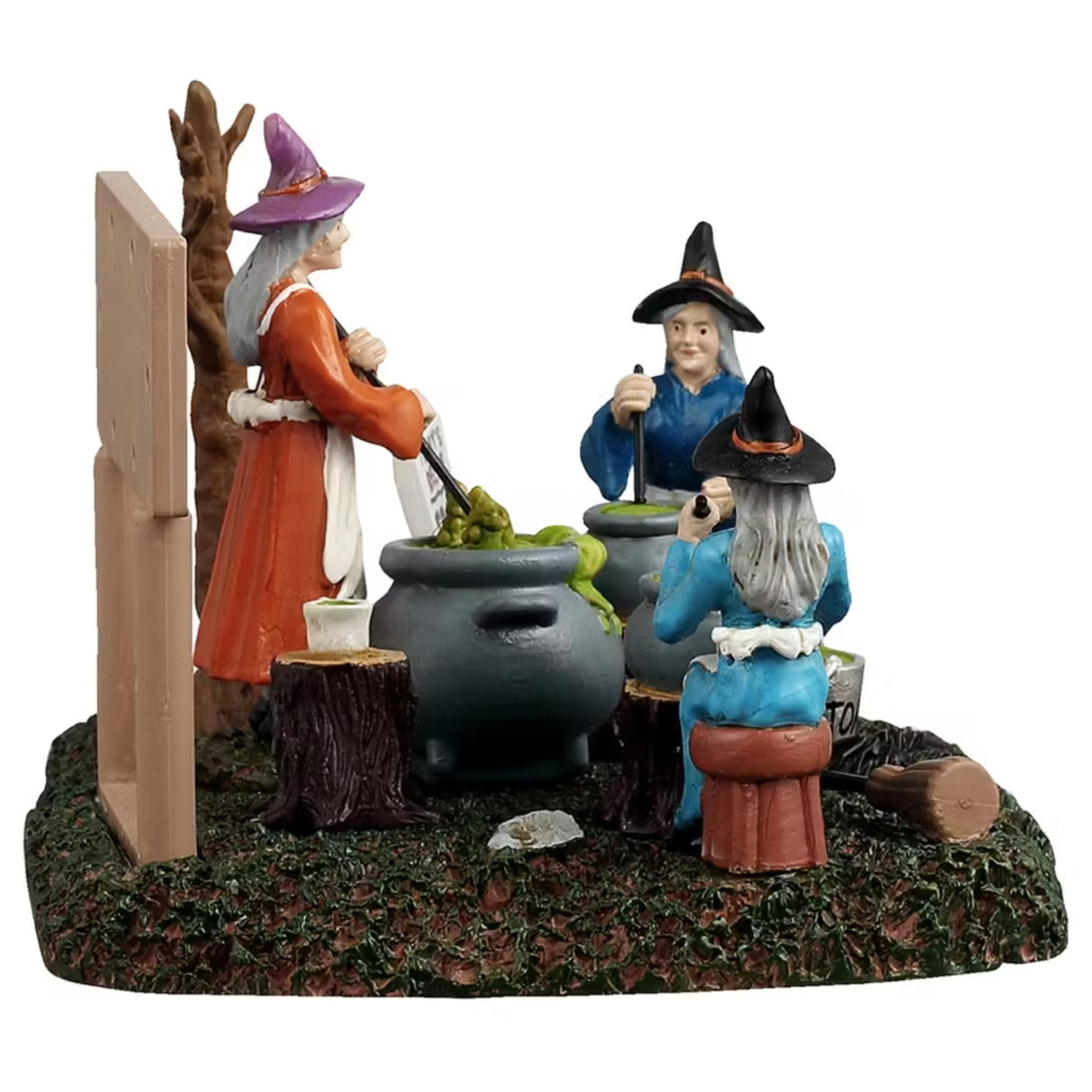 This is a different angle of the Lemax Spooky Town table accent designed to look like three witches sitting around stirring their individual cauldrons. There's a white board that reads "TOAD SOUP. Ingredients: 3 toads, 1 cup of swamp water, 1 cup of flies and 1 cup of worms." One witch is sitting on a stool and is wearing a blue cloak with a black hat. One witch is sitting on a tree stump and is wearing a dark blue cloak with a black hat. One witch is standing and is wearing an orange cloak with a pink hat.