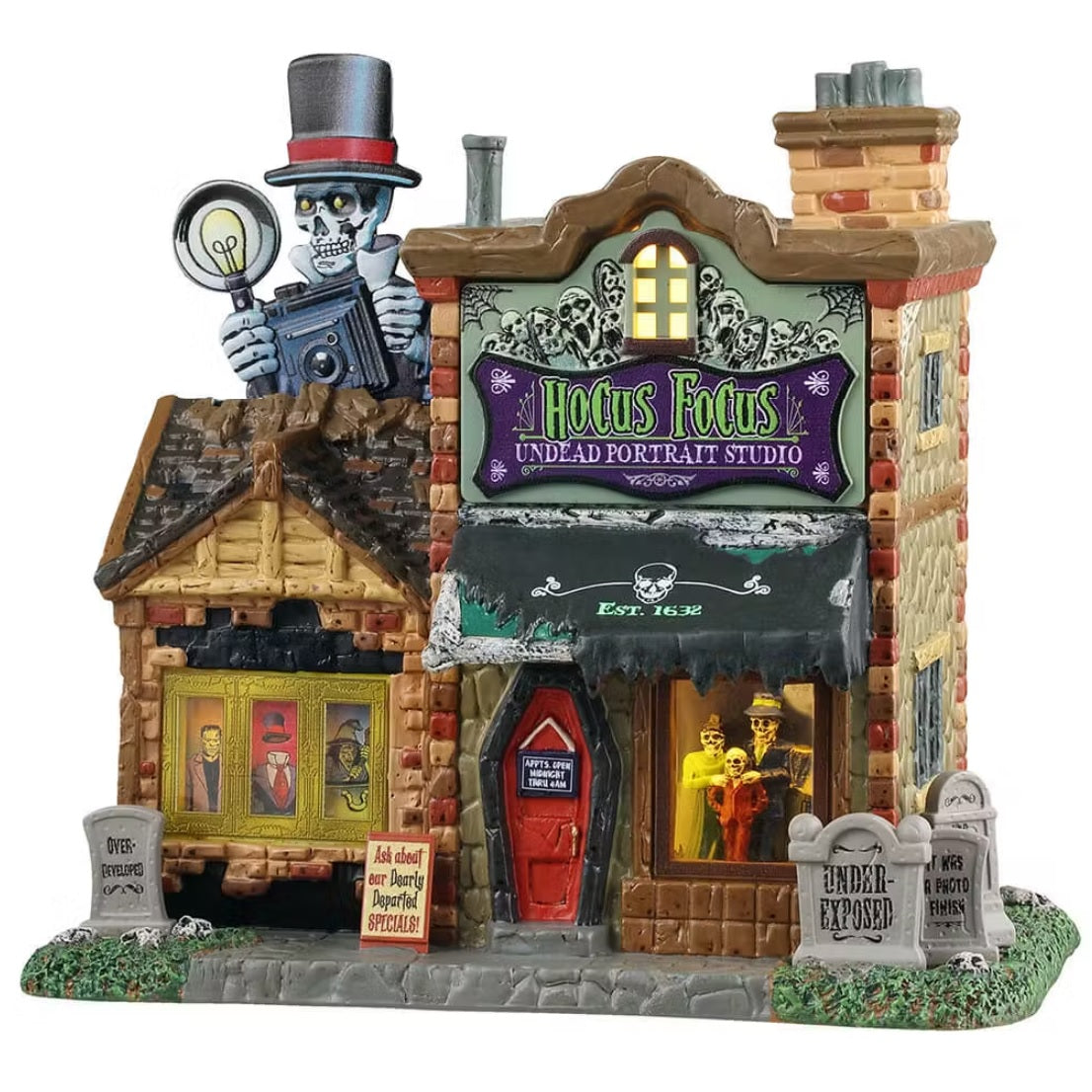 This is a Lemax Spooky Town building designed to look like an old brick building. There's a giant skeleton statue on the roof and it's wearing a top hat and holding an old-timey camera. The purple sign on the building reads "HOCUS FOCUS UNDEAD PORTRAIT STUDIO." Inside the windows, you can see monsters posing for pictures. On the sidewalk, there are tombstones and signs. The front door is shaped like a red coffin.