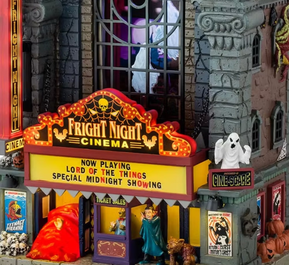 This is an up close view of this Lemax Spooky Town lighted building that's designed to look like a tall, gothic building that's been turned into a movie theater. It's called Fright Night Cinema. There are ghosts visible through the big window in the front. Underneath the marquee, there are skulls and a vampire standing next to a werewolf.
