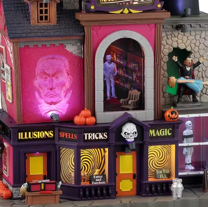 This is an up close view of this Smoke & Mirrors Magic Shop from Lemax. The windows on the bottom floor have mesmerizing yellow and black swirls on them. There's a big white skull above the front door. Off to the side, there's a magician making a woman float in the air.