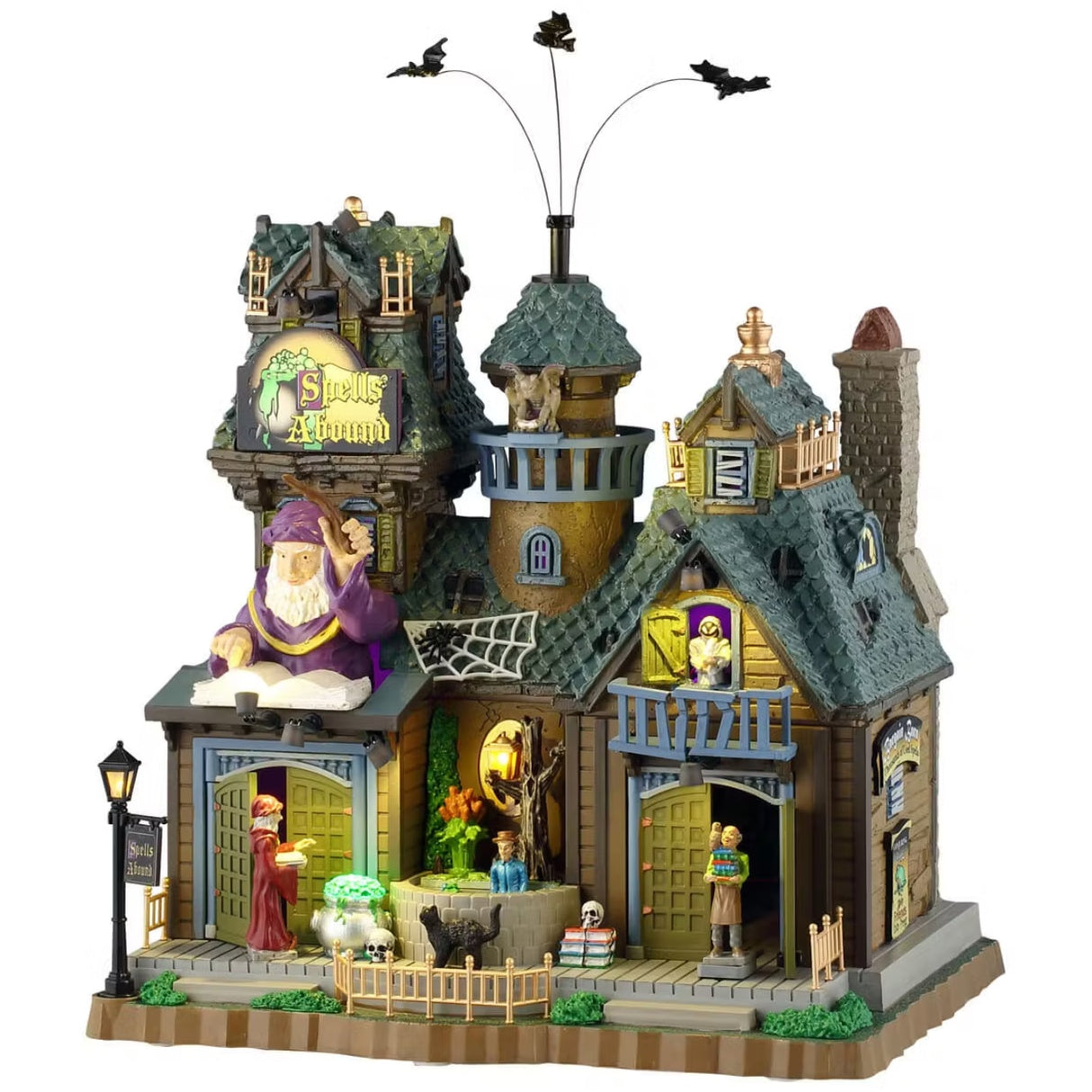 This is a Lemax Spooky Town building designed to look like a semi-rundown mansion. There's a sign that reads "SPELLS ABOUND." There are three bats flying above. There's a big wizard statue over one of the doors. You can see a wizard casting a spell on a cauldron, a person carrying books and a black cat on the sidewalk.