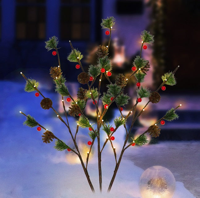 This is a lifestyle image showing the three branches staked into the snowy ground in front of a house. The warm white lights are on accenting the green pine needles, brown pine cones and red berries.