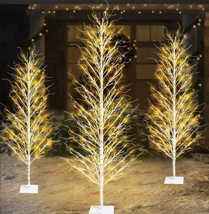 This is a lifestyle image showing the three different size options for this birch tree decoration. They're all set up on the snowy lawn in front of a house.