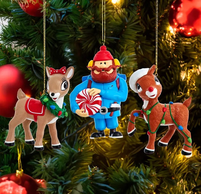 Ornaments designed to look like characters from the animated Christmas classic movie, Rudolph the Red Nosed Reindeer. One ornament is designed to look like Yukon Cornelius holding a peppermint. One ornament is designed to look like Clarice the Reindeer wearing a wreath. One ornament is designed to look like Rudolph the Reindeer wearing multi-colored Christmas lights and a Santa hat. This image shows what they look like against a lit Christmas tree.