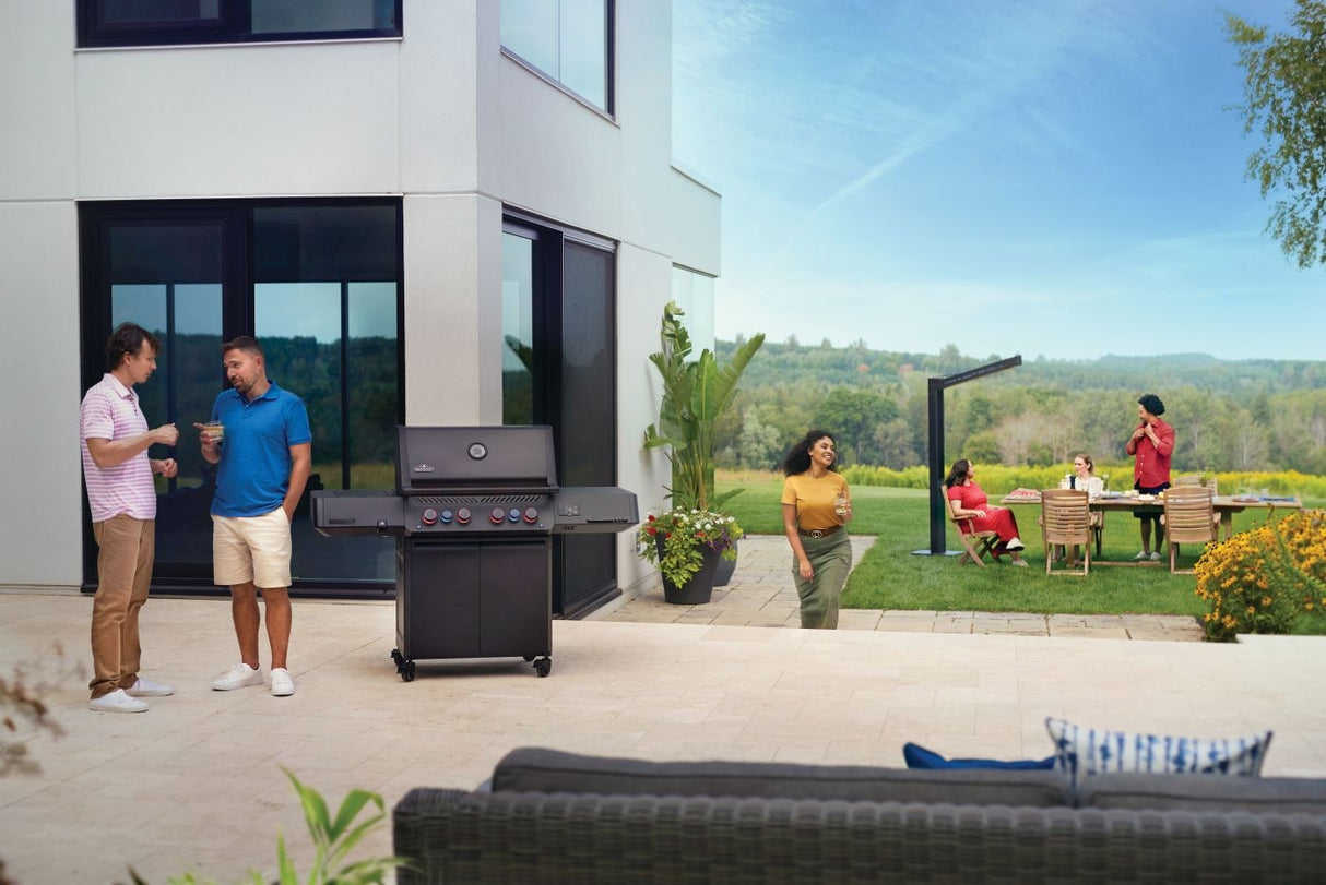 This is a lifestyle image for this specific grill. It's standing on a patio outside of a home. There are two men chatting on the side and there are people sitting at a table in the distance.