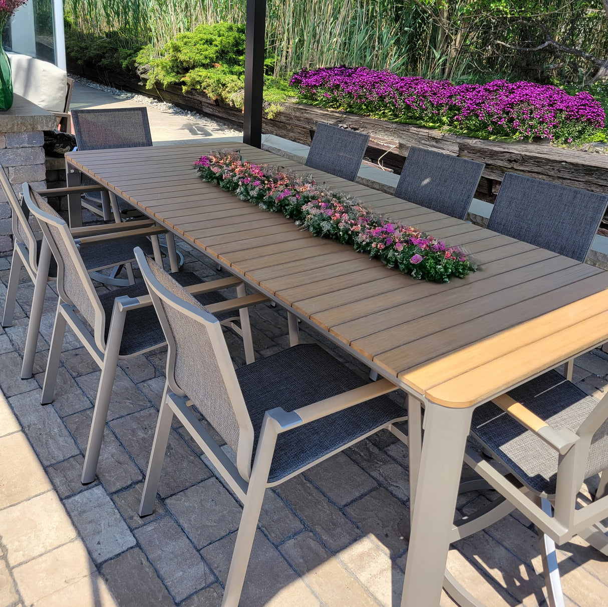 This image shows a patio dining set on a stone patio in front of a garden of beautiful, purple flowers. The frames of the chairs and the table are gray, the sling of the chairs is gray and the armrests and the top of the table are wood.