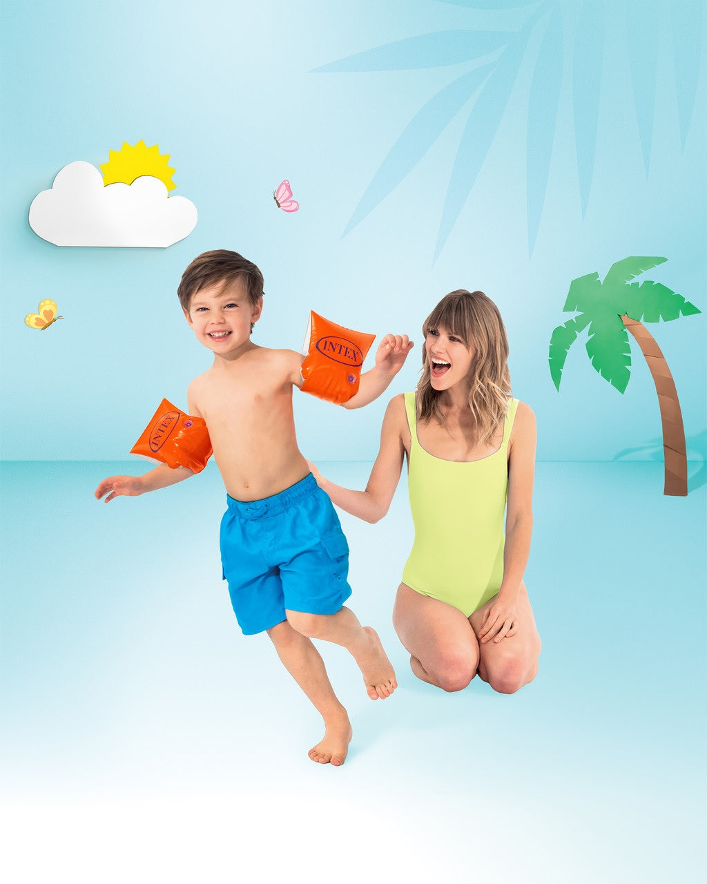 Neon orange colored inflatable arm bands for children. The inflatable goes around the two upper arm areas to help them stay afloat in water. This image appears to a photoshoot where the child and the mom are placed onto a cartoon looking backdrop. There's a sun in the sky, some butterflies and a palm tree and the people are designed to look like they're on the beach. The arm bands are around the small boy's arms.