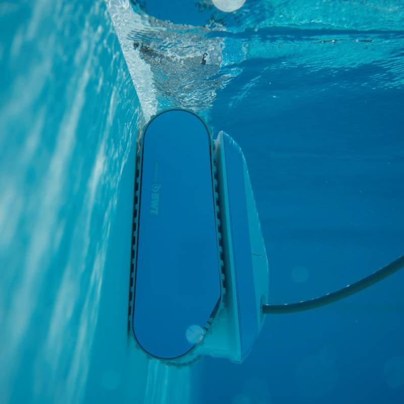 Robotic pool cleaner. Body is blue and white with gray bristles. This image shows the Cosmy vacuum underwater cleaning the walls of a pool.