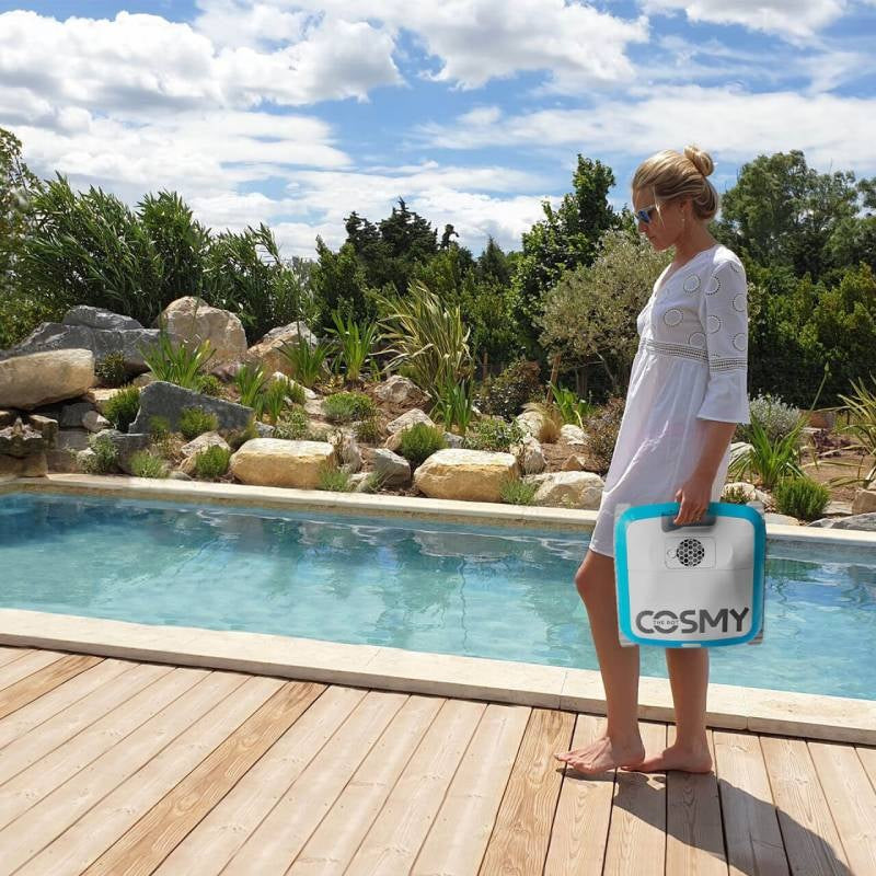 Robotic pool cleaner. Body is blue and white with gray bristles. This picture shows a woman holding the Cosmy vacuum next to a pool on a sunny day.