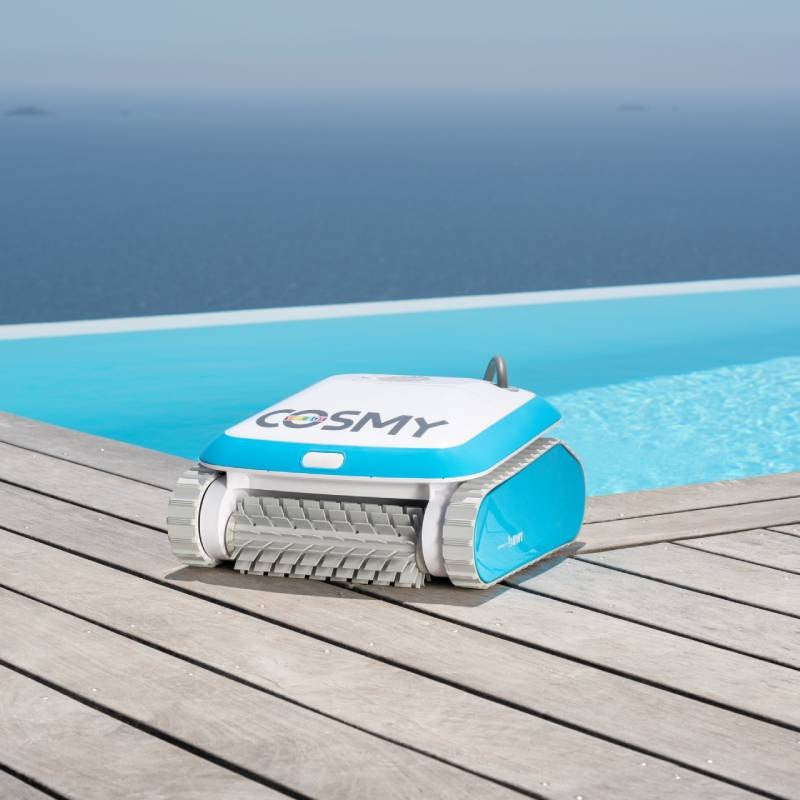 Robotic pool cleaner. Body is blue and white with gray bristles. This image shows the Cosmy vacuum sitting on a deck next to an inground pool.