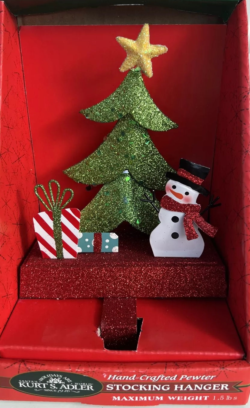 Stocking hanger covered in glitter. Designed to look like a light green tree with a yellow star on top. Next to the tree are two presents and a snowman wearing a top hat and a red scarf. The base of the holder is covered in red glitter.