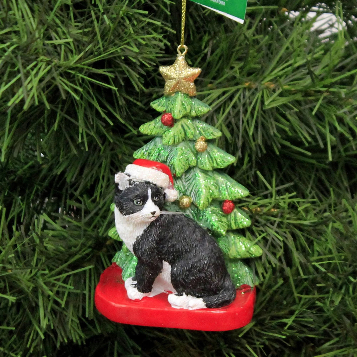 Ornaments shaped like a cat in front of a tree. One ornament features a black and white cat. One ornament features an orange and white cat. One ornament features a light gray and dark gray cat. One ornament features a white cat. All the cats are wearing Santa hats.