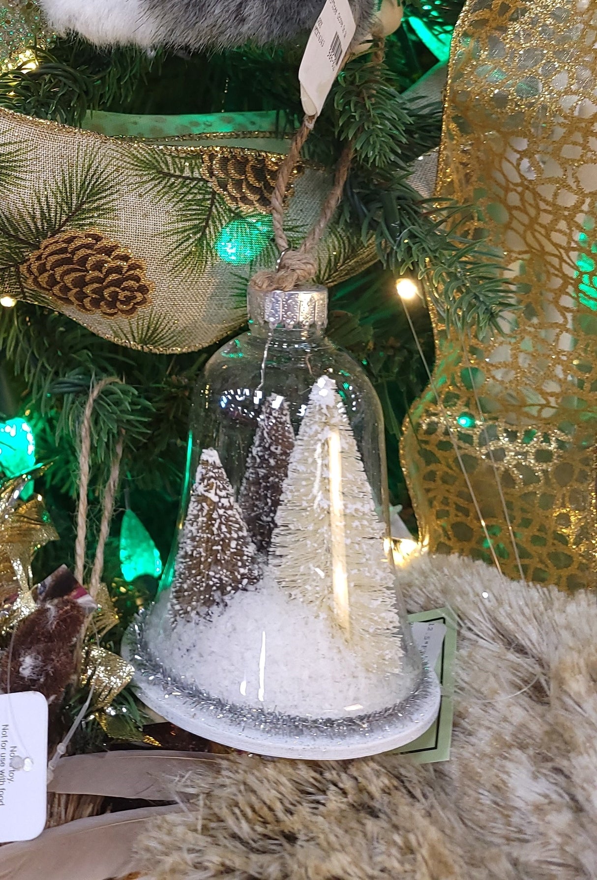 Ornament shaped like a glass bell with small bottle brush trees inside. One ornament features two green trees and one yellow tree. One ornament features two reddish brown trees and one yellow tree. All the trees are designed to appear covered in snow. Each ornament features tinsel around the border.