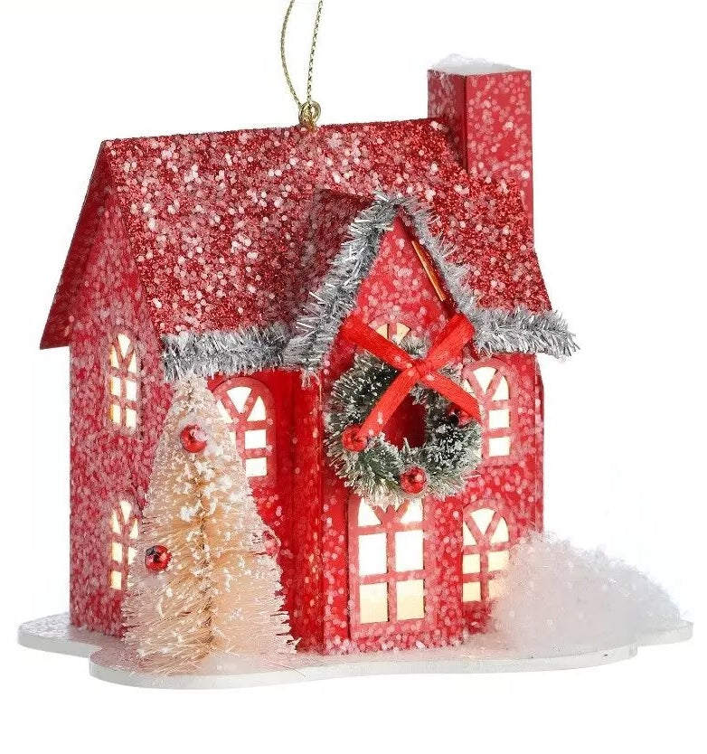 Ornament shaped like a house covered in snow. One house is red and features a wreath near the roof with a red bow. One house is green and features a wreath near the roof with a green bow.