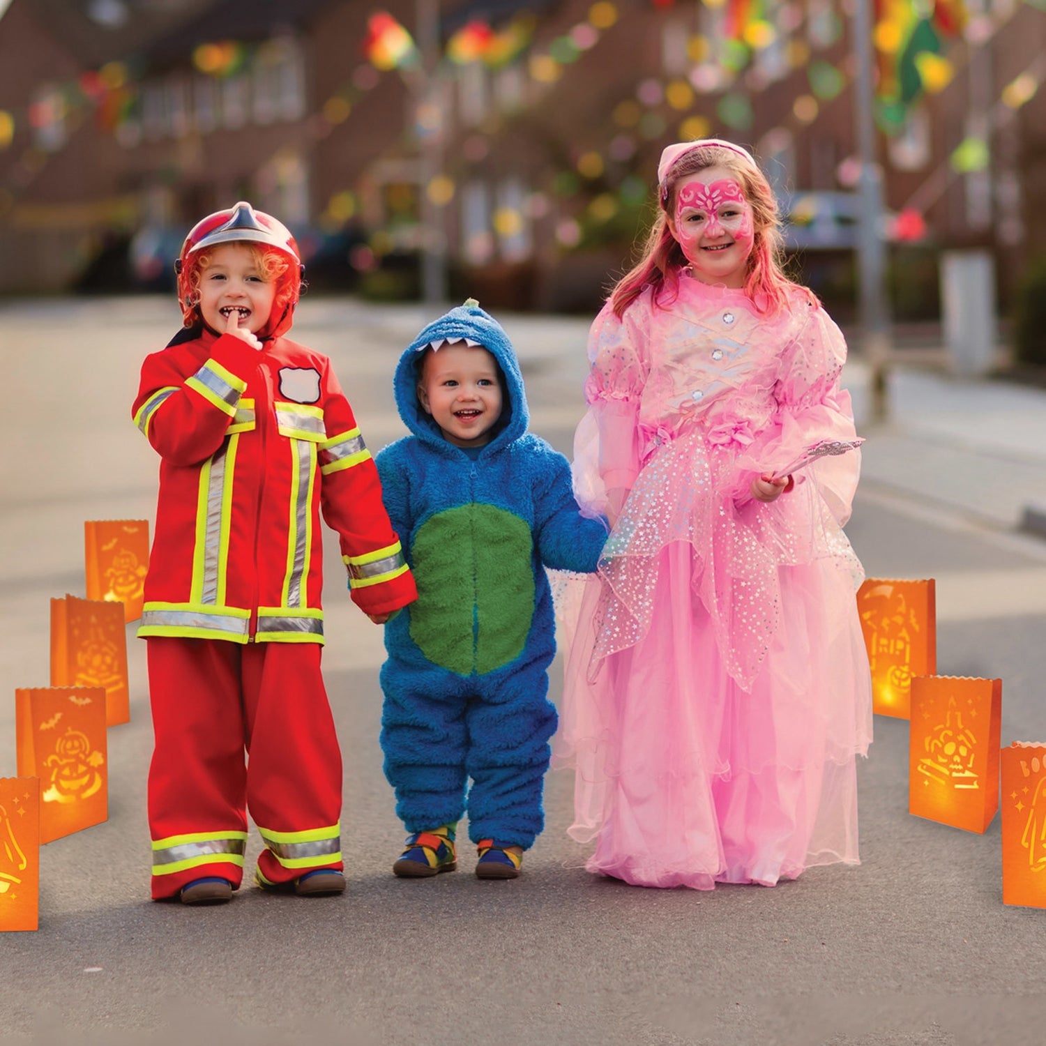 This is a lifestyle image showing these luminary bags in use. This picture shows three children walking along a pathway lined with them. One child is dressed like a firefighter. One child is dressed like a blue monster. The third child is dressed like a pink princess.
