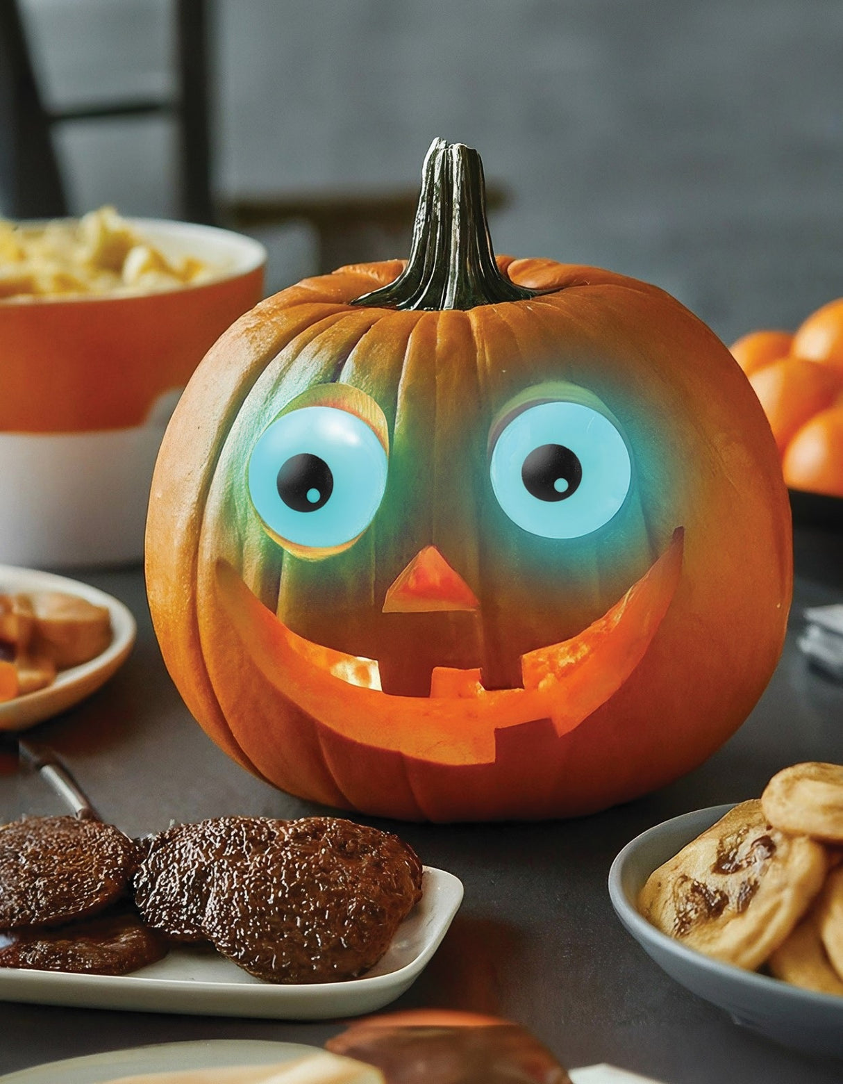 This is a lifestyle image showing the eyeballs in use. They're inside a pumpkin, glowing blue. The pumpkin has a carved nose and a carved smile and is in the center of a table full of food.