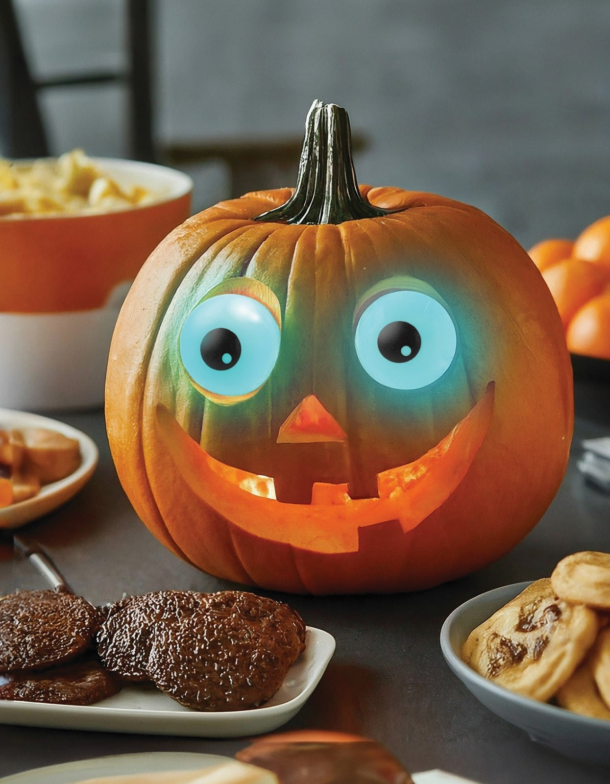 This is a lifestyle image showing the eyeballs in use. They're inside a pumpkin, glowing blue. The pumpkin has a carved nose and a carved smile and is in the center of a table full of food.