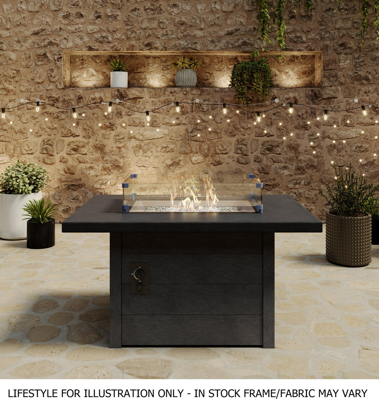A rectangular fire pit table on an outdoor stone porch with a stone wall and surrounded by plants
