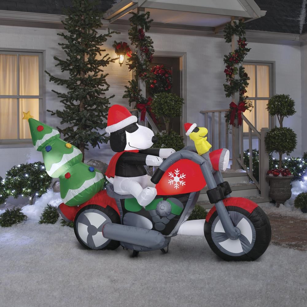 Christmas inflatable. It features Snoopy wearing a Santa hat and wearing sunglasses. Snoopy is driving a red motorcycle with a snowflake accent on it. There's a decorated Christmas tree on the back and Woodstock is riding on the handlebars. There are LED lights inside.