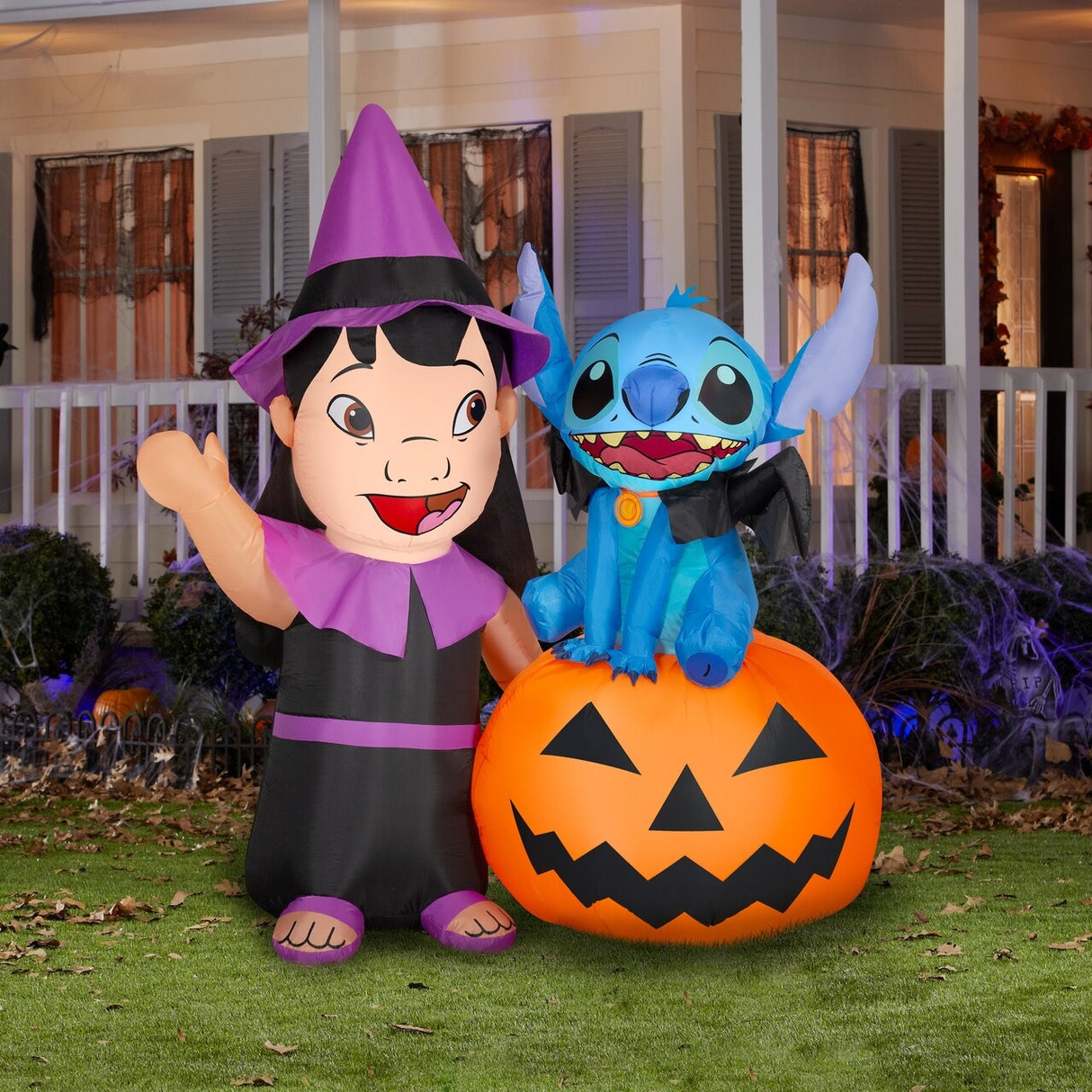 Halloween inflatable shaped like the Disney characters Lilo and Stitch dressed for Halloween. Lilo is wearing a witch costume and Stitch is wearing a collar with bat wings on it. Stitch is sitting on a jack-o-lantern.