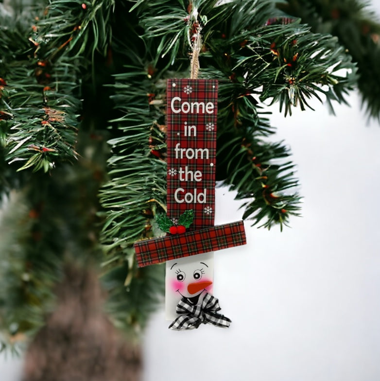 Hanging sign designed to look like a snowman head. One snowman has a red and green buffalo check top hat that reads "Let It Snow." One snowman has a red and green plaid top hat that reads "Come in from the Cold." One snowman has a red and white plaid top hat that reads "There's Snow place like Home."