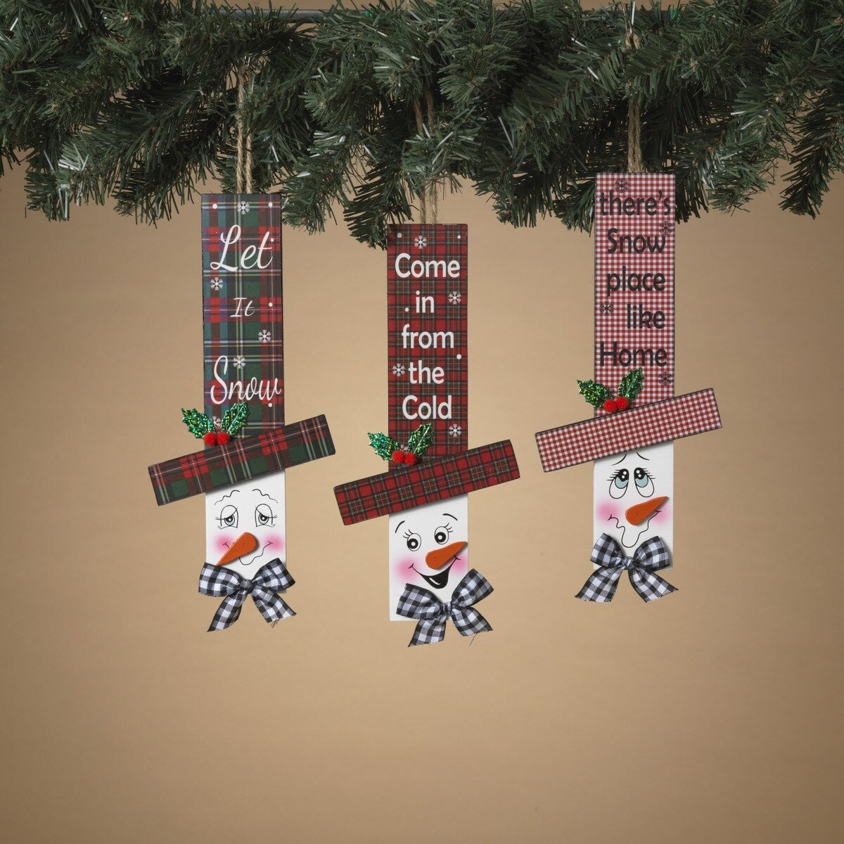 Hanging sign designed to look like a snowman head. One snowman has a red and green buffalo check top hat that reads "Let It Snow." One snowman has a red and green plaid top hat that reads "Come in from the Cold." One snowman has a red and white plaid top hat that reads "There's Snow place like Home."