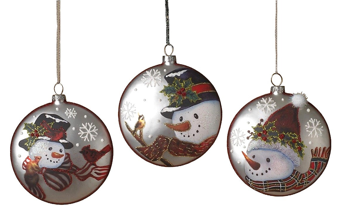 Round ornament featuring a snowman in a winter scene. One ornament has a snowman wearing a red and white scarf and a top hat and is smiling at a squirrel and two cardinals. One ornament has a snowman wearing a red and gold scarf and a top hat and is smiling at a bird on a branch. One ornament has a snowman wearing a red, green and gold scarf and a Santa hat. Each ornament features holly leaves and berries and snowflakes.