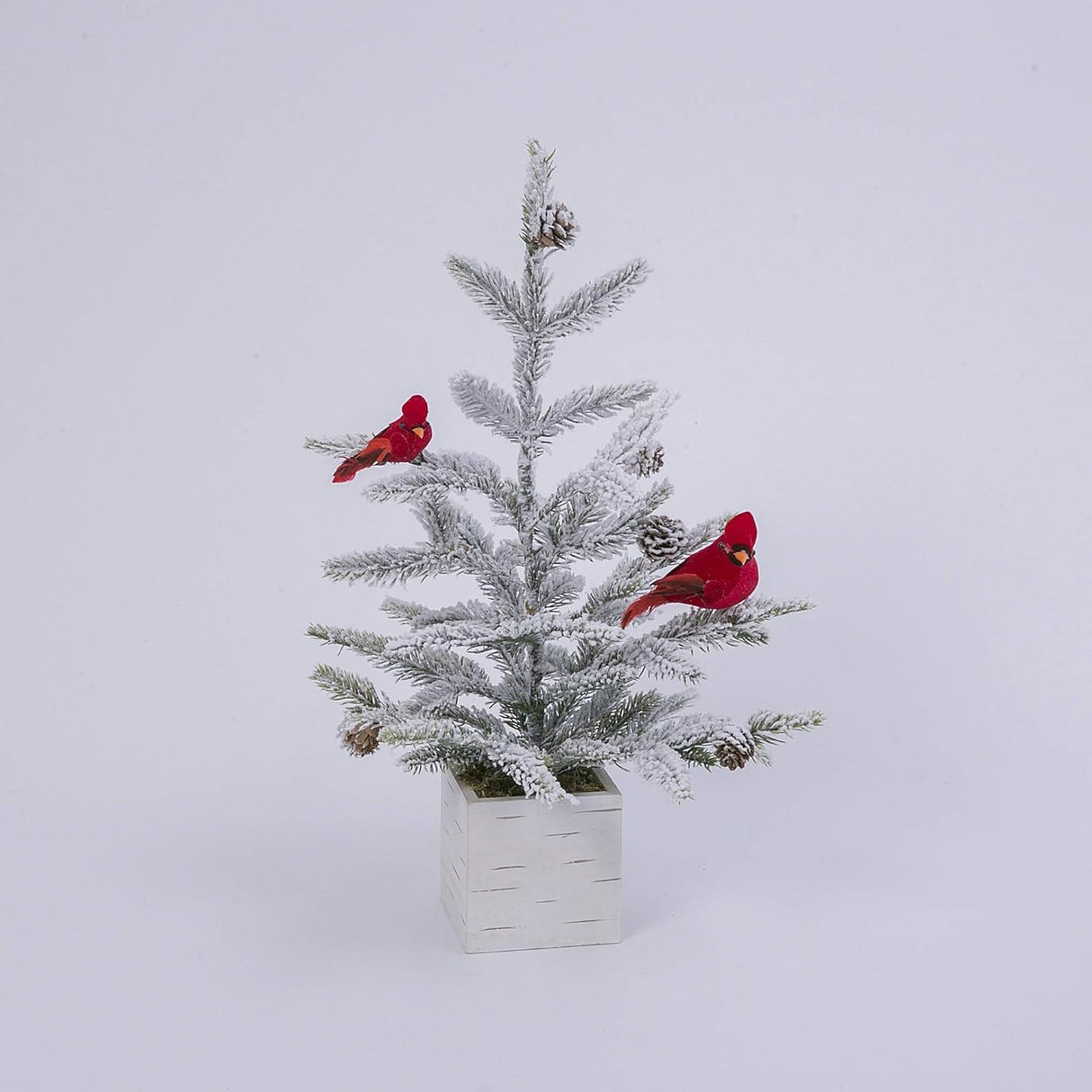 Tabletop tree in a birch planter box. The branches of the tree are dusted with white to mimic snow. The tree is accented with two red cardinal birds and some snow-covered pine cones.
