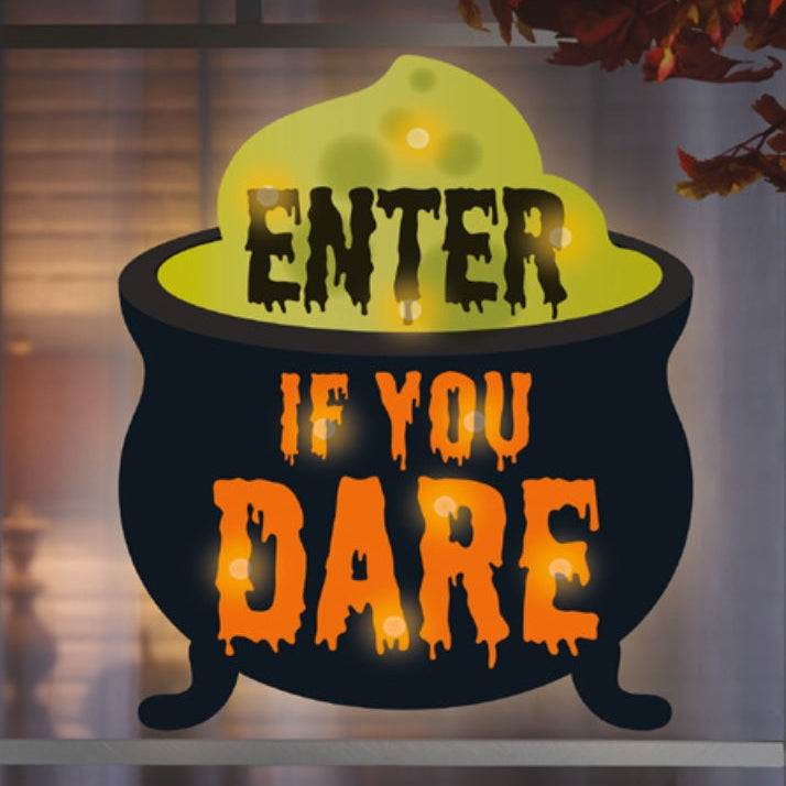This is a lifestyle image showing this Halloween decoration hanging in a window. It's designed to look like a black cauldron with yellow goo coming out of it. It reads "Enter if you dare."