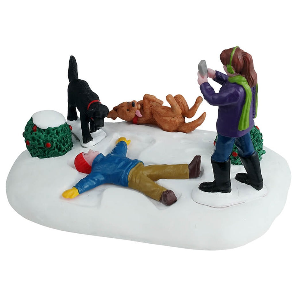 Resin table accent for a Lemax Village tabletop display. Designed to look like a young child making a snow angel while a girl takes a picture of the scene. There are two dogs and two bushes nearby. One dog is black and one is a golden brown color. The golden brown dog is laying on it's back with it's tongue hanging out. The ground is covered in snow and both kids are wearing winter clothes.