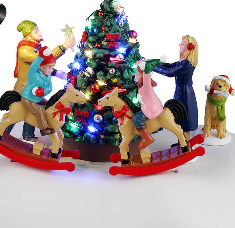 Animated resin village accessory for a Lemax display. Designed to look like a family decorating a Christmas tree on a snowy patch. They're all wearing winter clothes. The kids are riding on reindeer that rock back and forth. There's also a dog watching, wearing a Santa hat. There's a pole nearby with a light on it.