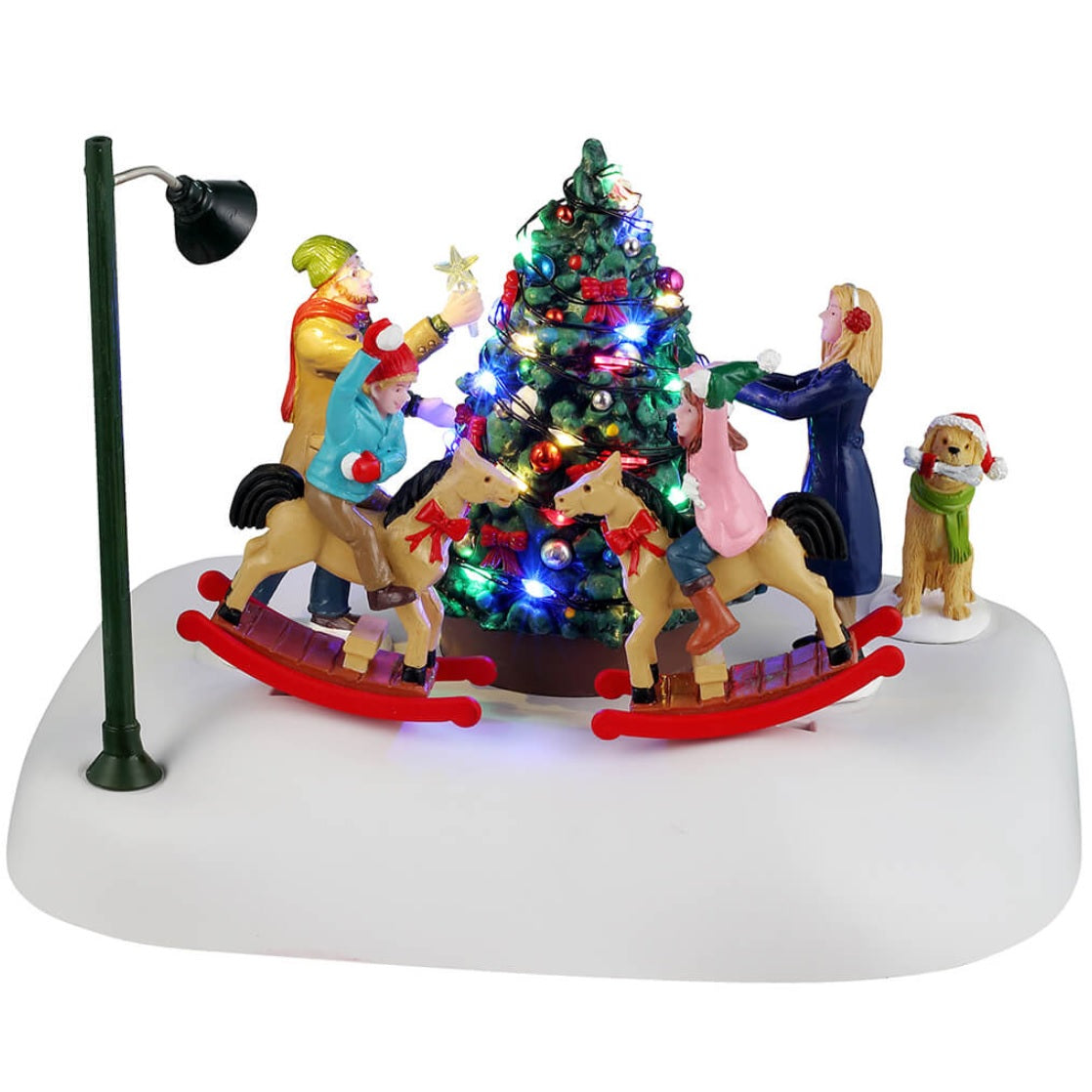 Animated resin village accessory for a Lemax display. Designed to look like a family decorating a Christmas tree on a snowy patch. They're all wearing winter clothes. The kids are riding on reindeer that rock back and forth. There's also a dog watching, wearing a Santa hat. There's a pole nearby with a light on it.