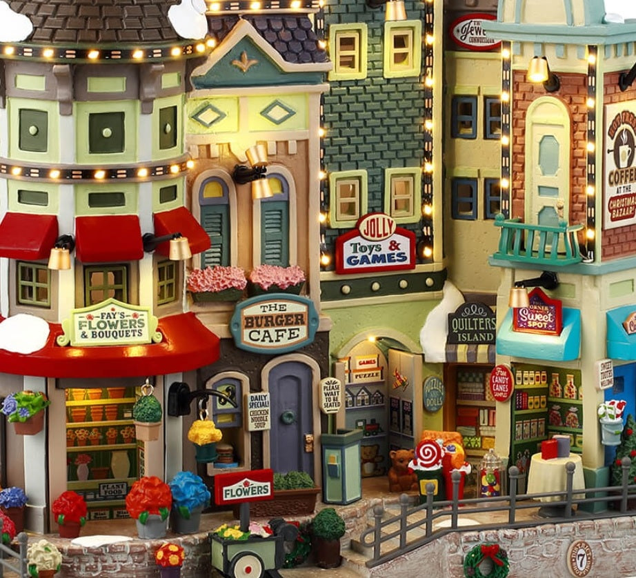 Resin building facade for a Lemax display. Designed to look like a bunch of store fronts. Signs read: "Fay's Flowers & Bouquets," "The Burger Cafe," "Jolly Toys & Games," "Quilter's Island," Sweet Spot" and "Hot Fresh Coffee at the Christmas Bazaar!" There are flowers everywhere and a billboard on the roof advertising a visit with Santa.