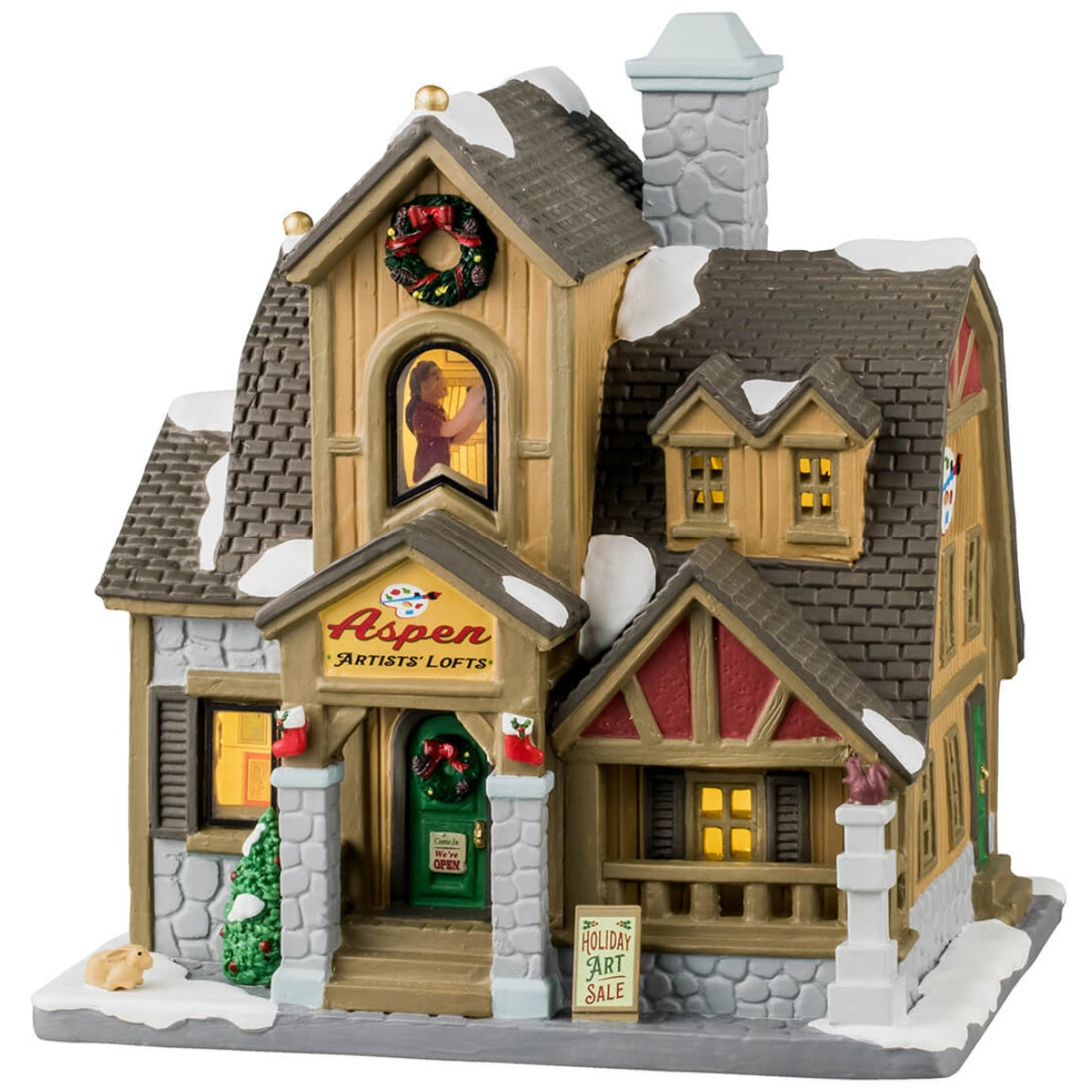 Porcelain lighted building for a Lemax display. Designed to look like a charming house covered in snow. There's a decorated wreath near the roof and on the front door. There's a decorated tree outside and stockings hanging on the sides of the entrance. You can see someone painting inside. The sign on the building reads "Aspen Artist's Lofts" and the sign outside the porch reads "Holiday Art Sale!" There's a cute bunny in the snow and a squirrel on a column near the porch.