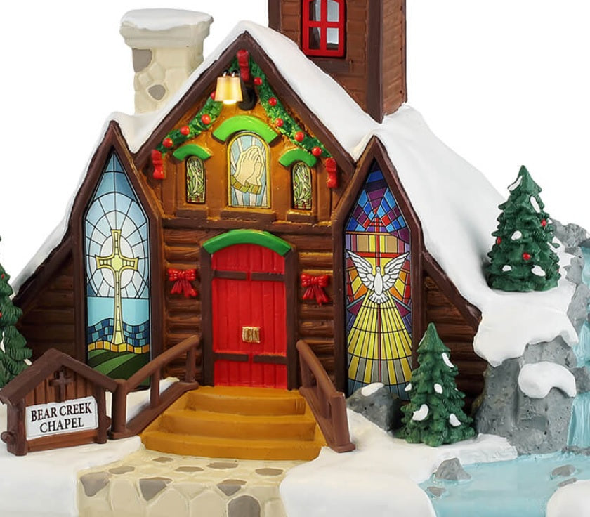 Resin lighted building for a Lemax display. Designed to look like a brown, wooden chapel with a stone chimney and a bell tower. The roof and the ground is covered in snow. There's a wreath on the bell tower, garland on the roof and two red bows on the sides of the door. The windows are stained glass. One of them is a yellow cross with a blue background. The other is a white dove with a yellow glow. There's a waterfall on the side.