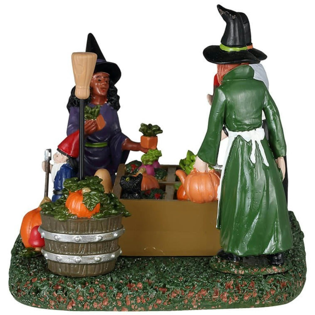 Resin table piece for Lemax display. Designed to look like three witches tending to a garden. There are pumpkins and radishes and lettuce and mushrooms and a black cat surveying the plants. There's a gnome statue holding a shovel.