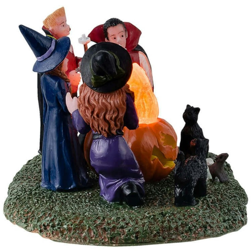 Resin table accent for a Lemax display. Designed to look like four kids dressed in costumes sitting around a fire roasting marshmallows. Two of the kids are dressed like witches and the other two are dressed like vampires. There are black cats and rats watching.