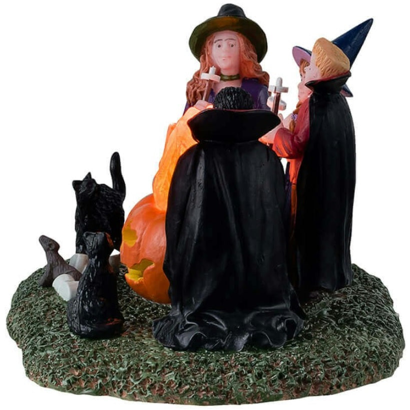 Resin table accent for a Lemax display. Designed to look like four kids dressed in costumes sitting around a fire roasting marshmallows. Two of the kids are dressed like witches and the other two are dressed like vampires. There are black cats and rats watching.