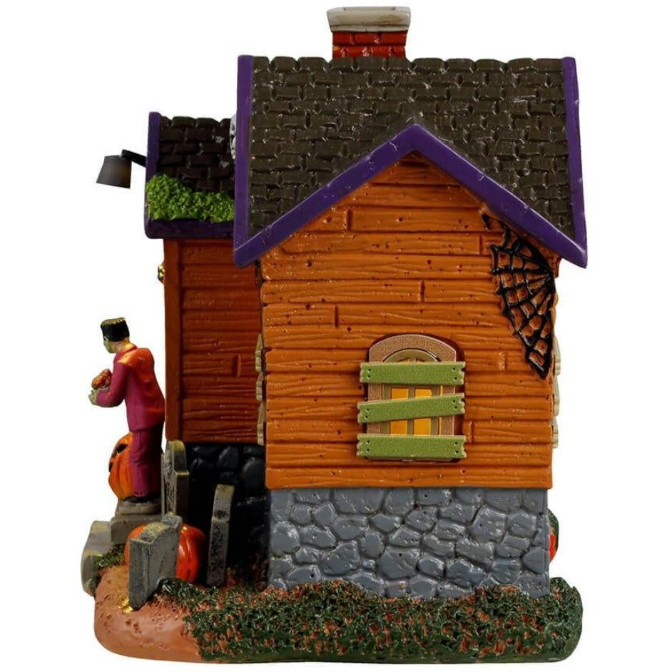 Resin building for a Lemax display. Designed to look like an orange, rundown cabin with boarded up windows and spiders everywhere. Frankenstein is standing on the porch. There are tombstones and carved pumpkins on the front lawn.