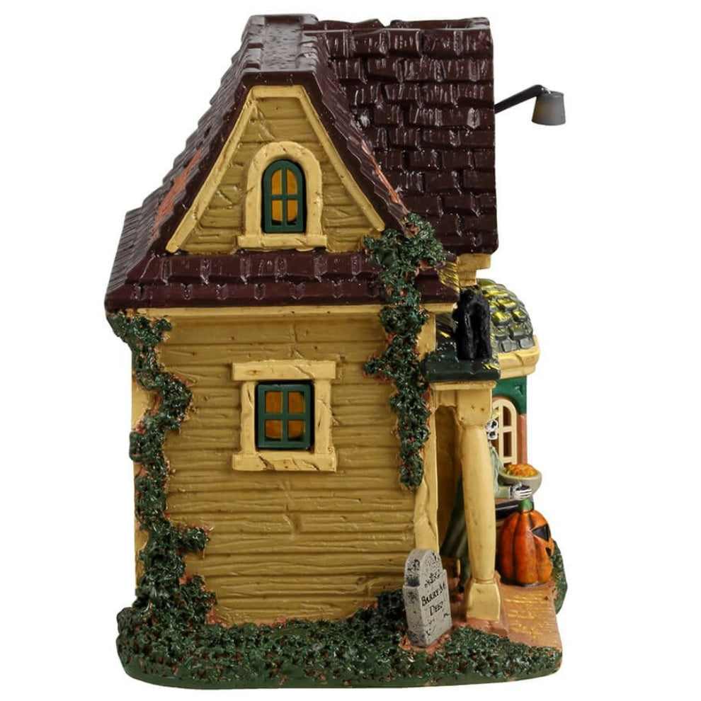 Resin building for a Lemax display. Designed to look like a run down house with moss growing on the side. Part of the roof is missing. There's a black cat on the roof and spiders. There's a skeleton wearing a lime green suit and cape handing out candy. There are tombstones in front of the house and a carved jack-o-lantern.