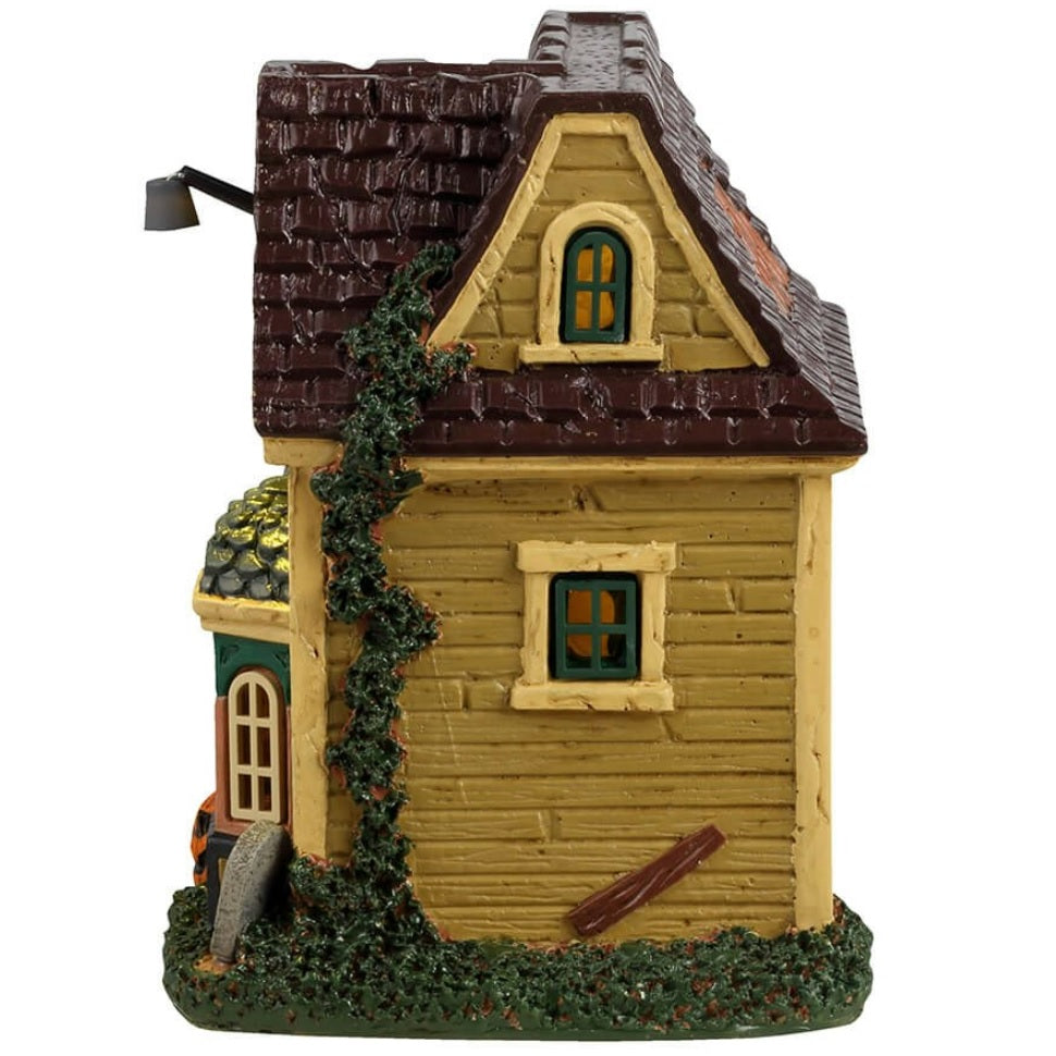 Resin building for a Lemax display. Designed to look like a run down house with moss growing on the side. Part of the roof is missing. There's a black cat on the roof and spiders. There's a skeleton wearing a lime green suit and cape handing out candy. There are tombstones in front of the house and a carved jack-o-lantern.