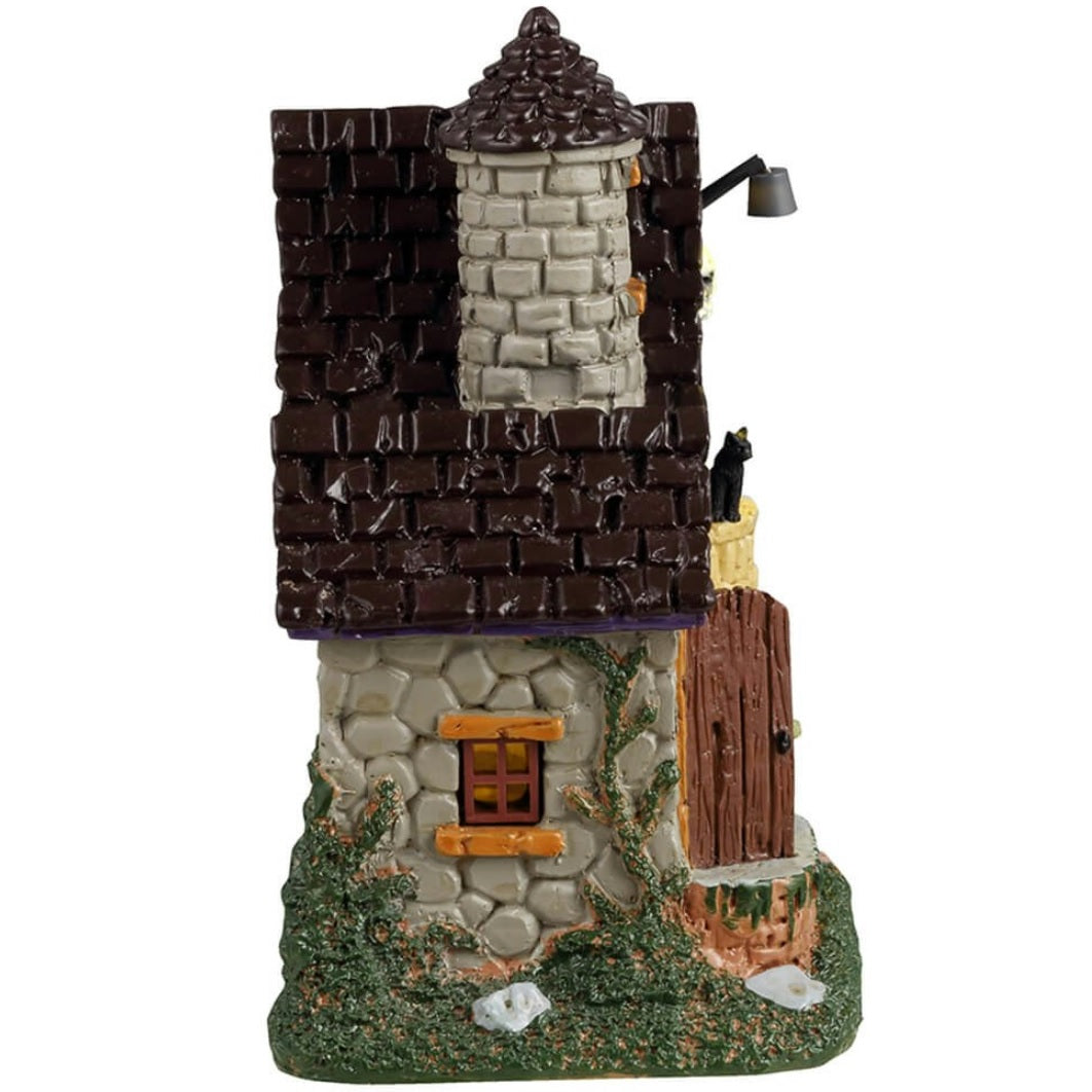 Resin building for a Lemax display. Designed to look like a stone house with moss growing on the side. There's a witch handing out candy at the front door and a skeleton sitting on the ground next to a pumpkin.