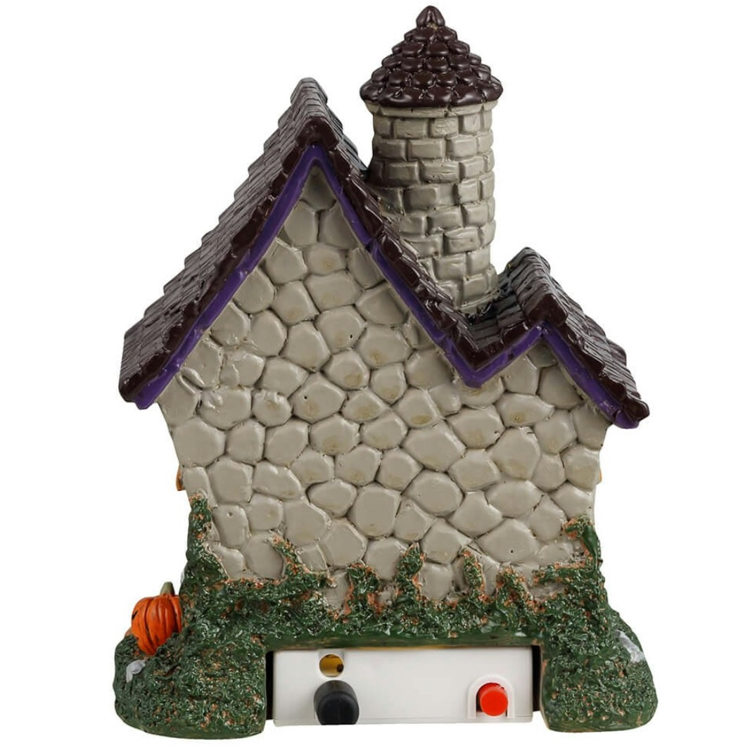 Resin building for a Lemax display. Designed to look like a stone house with moss growing on the side. There's a witch handing out candy at the front door and a skeleton sitting on the ground next to a pumpkin.