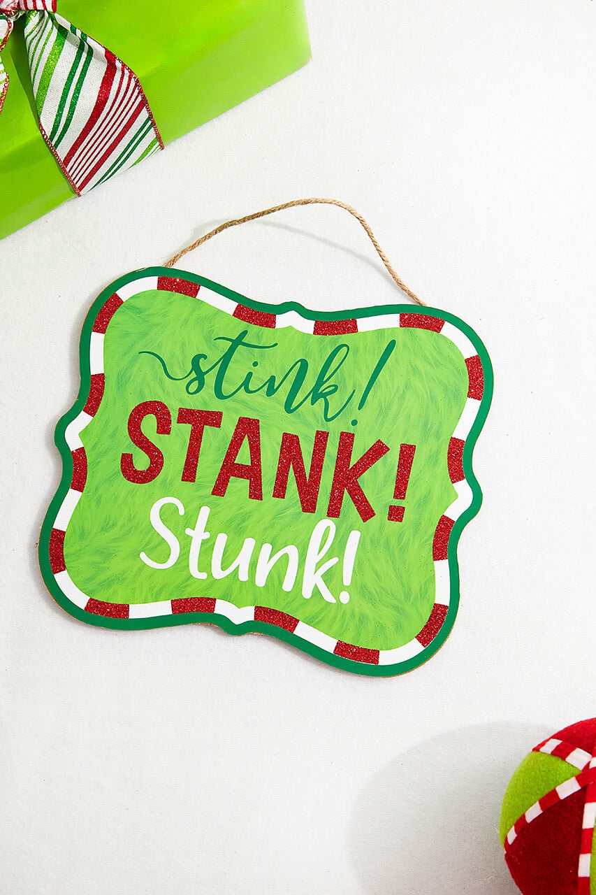 Hanging holiday sign that reads "stink! STANK! Stunk!" in a green, red glitter and white font. The background is designed to look like green fur. There's a red and white border up against a solid green border. Hangs from a jute rope.